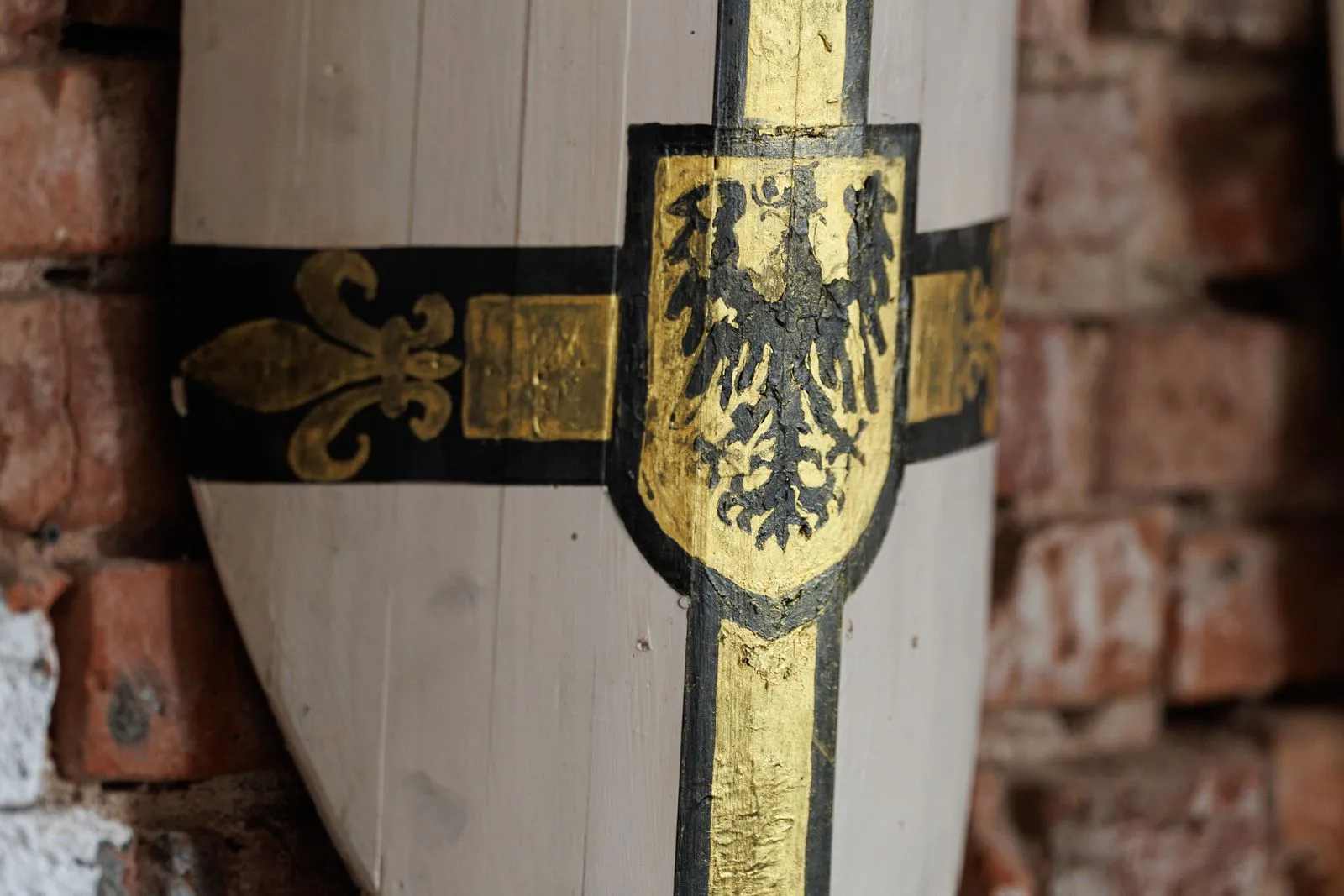 Close-up of a white wooden shield with a black and gold cross, a black eagle on a gold crest and gold fleurs-de-lis, leaning against a weathered red brick wall