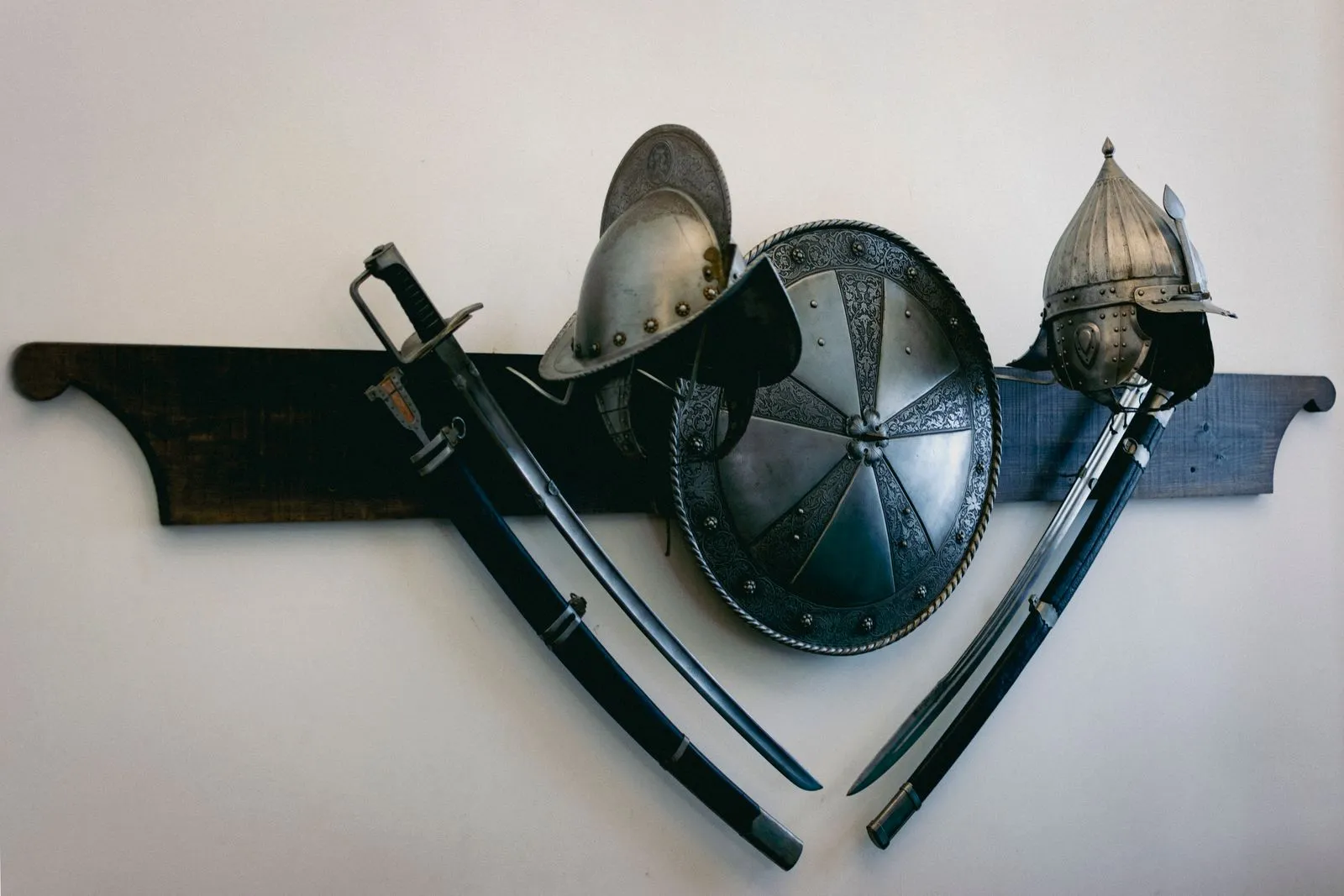 Escudo redondo de metal gravado, dois elmos e dois sabres cruzados presos a uma tábua de madeira numa parede branca de museu